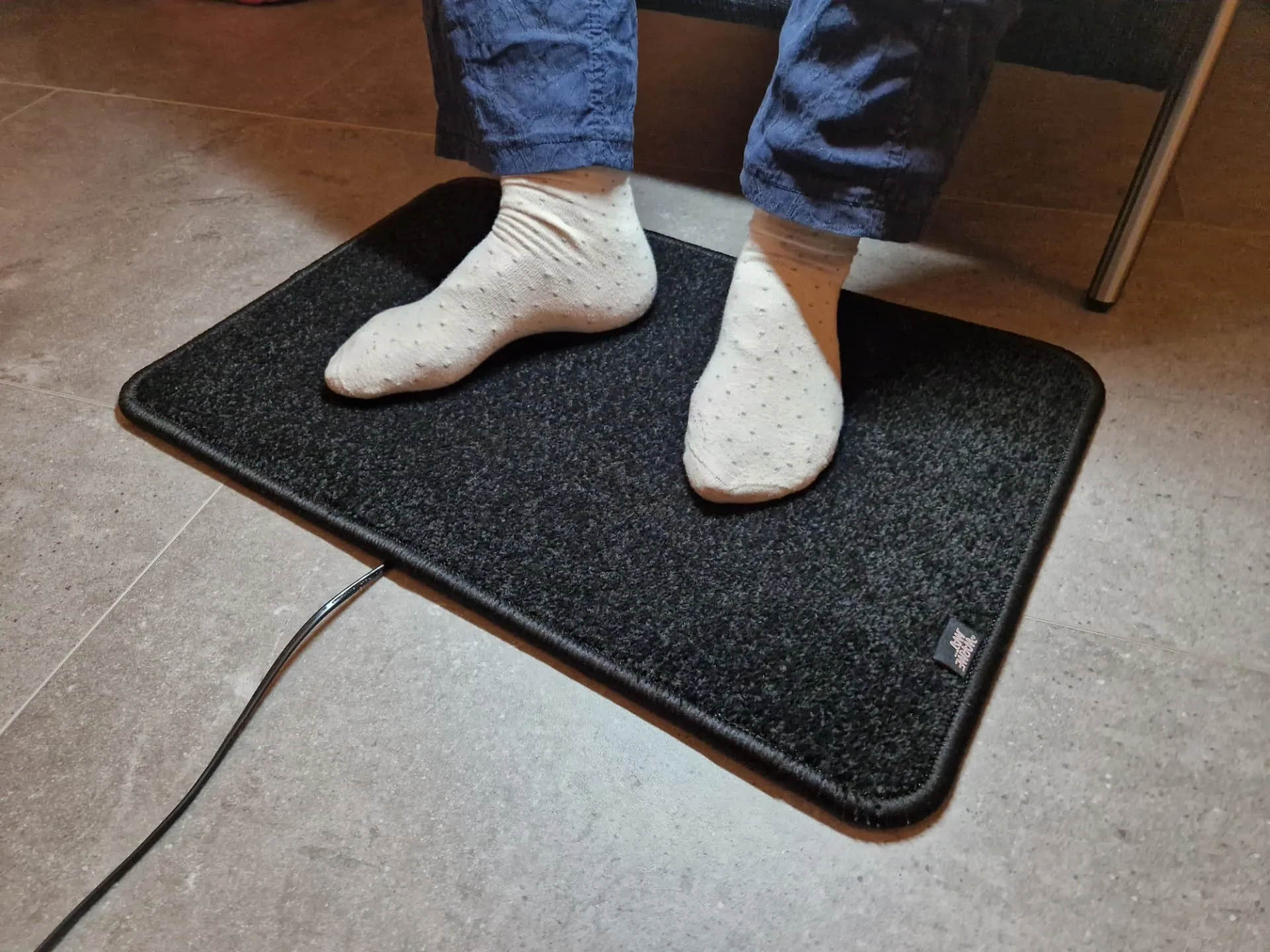 Zwarte verwarmde voetenmat Excellent met voeten in sokken op een lichte tegelvloer, met het De WarmeMat-label op de rand